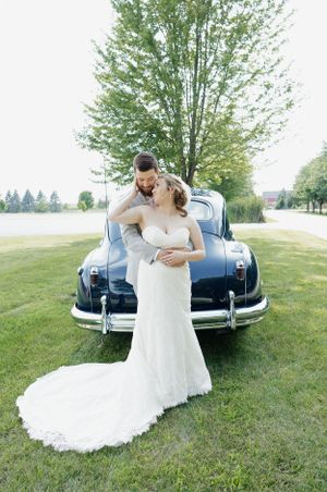 Romantic wedding couple posing with vintage blue car in countryside setting.