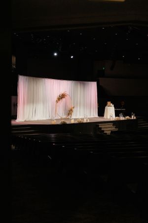 Wedding ceremony stage setup with white draping and gold arch in a dark auditorium.
