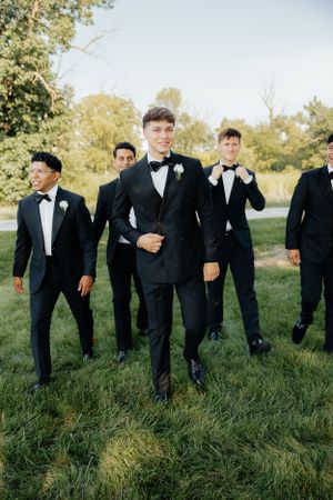 Wedding party in black tuxedos posing on grass during outdoor photo session.