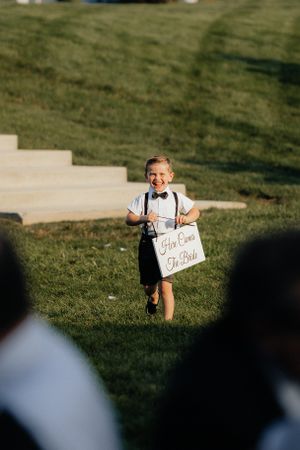 Young ring bearer holds a handwritten sign while walking down a grassy aisle at an outdoor wedding ceremony.
