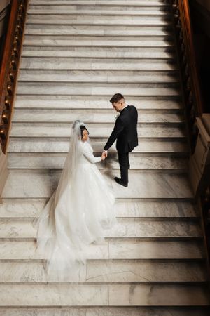 A bride and groom walk up grand marble stairs with her dress trailing behind.