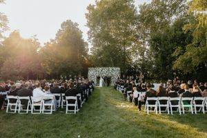 Rows of white chairs line a sunlit outdoor wedding ceremony space with trees in the background.
