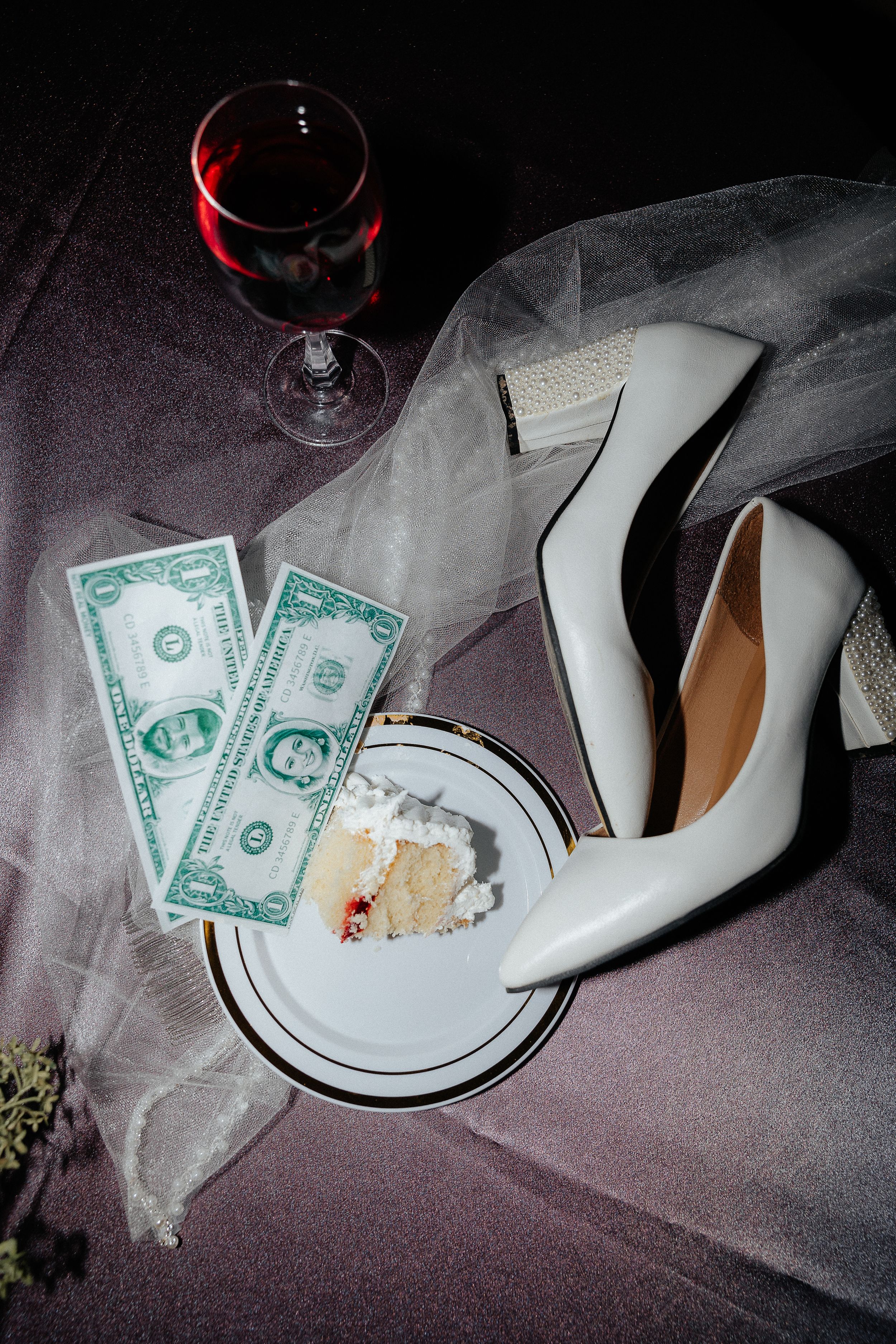 Wedding details including white heels, money, wine glass and cake on a dark surface.