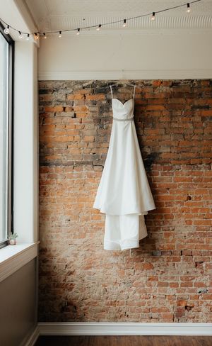 White wedding dress hanging against exposed brick wall with string lights creating romantic ambiance.