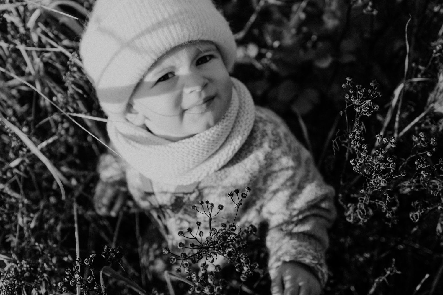 A joyful child wearing a winter hat and scarf sits among greenery in black and white photography.