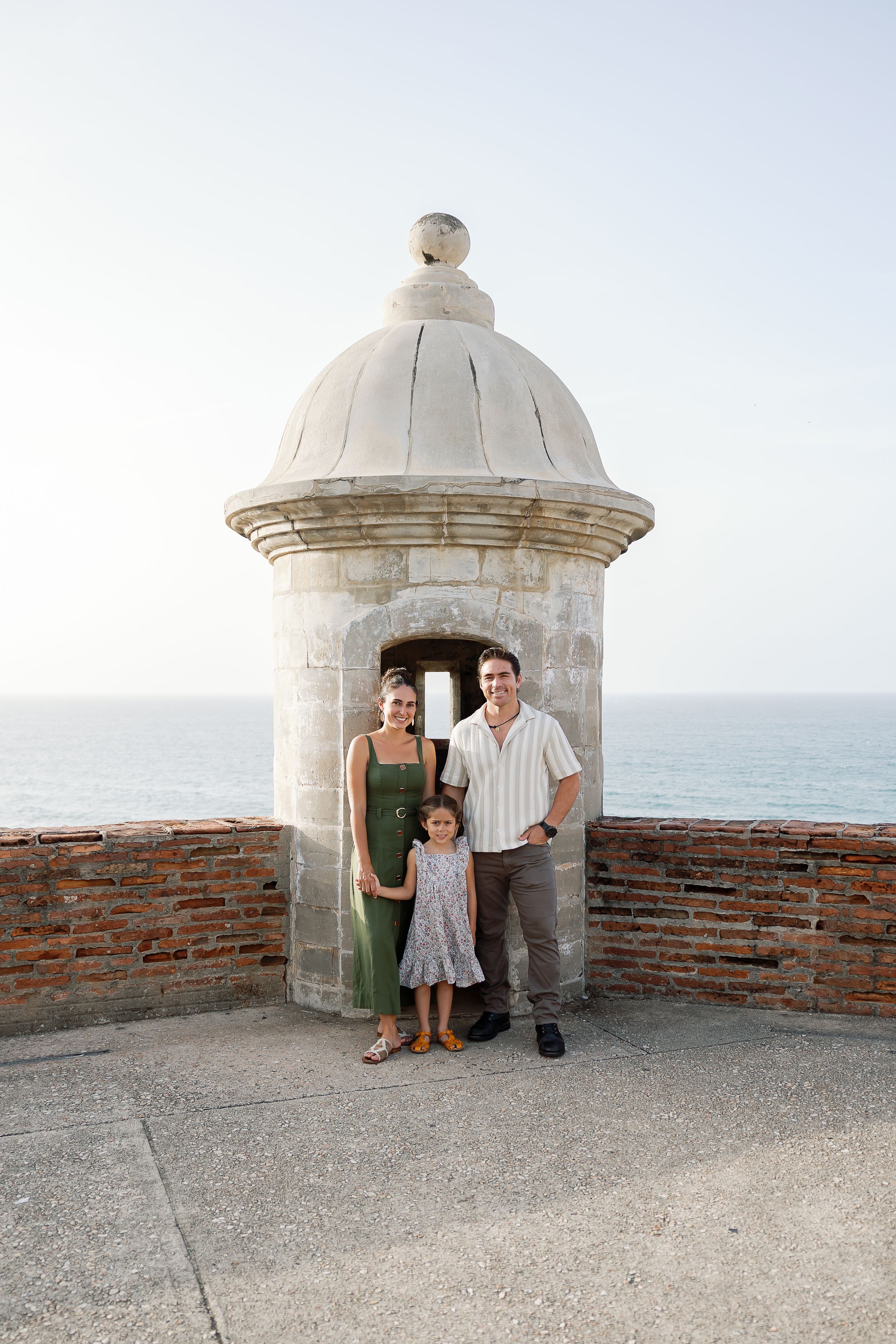 A family of three, a mother, father, and young child, standing together in front of a historic domed building overlooking the ocean.