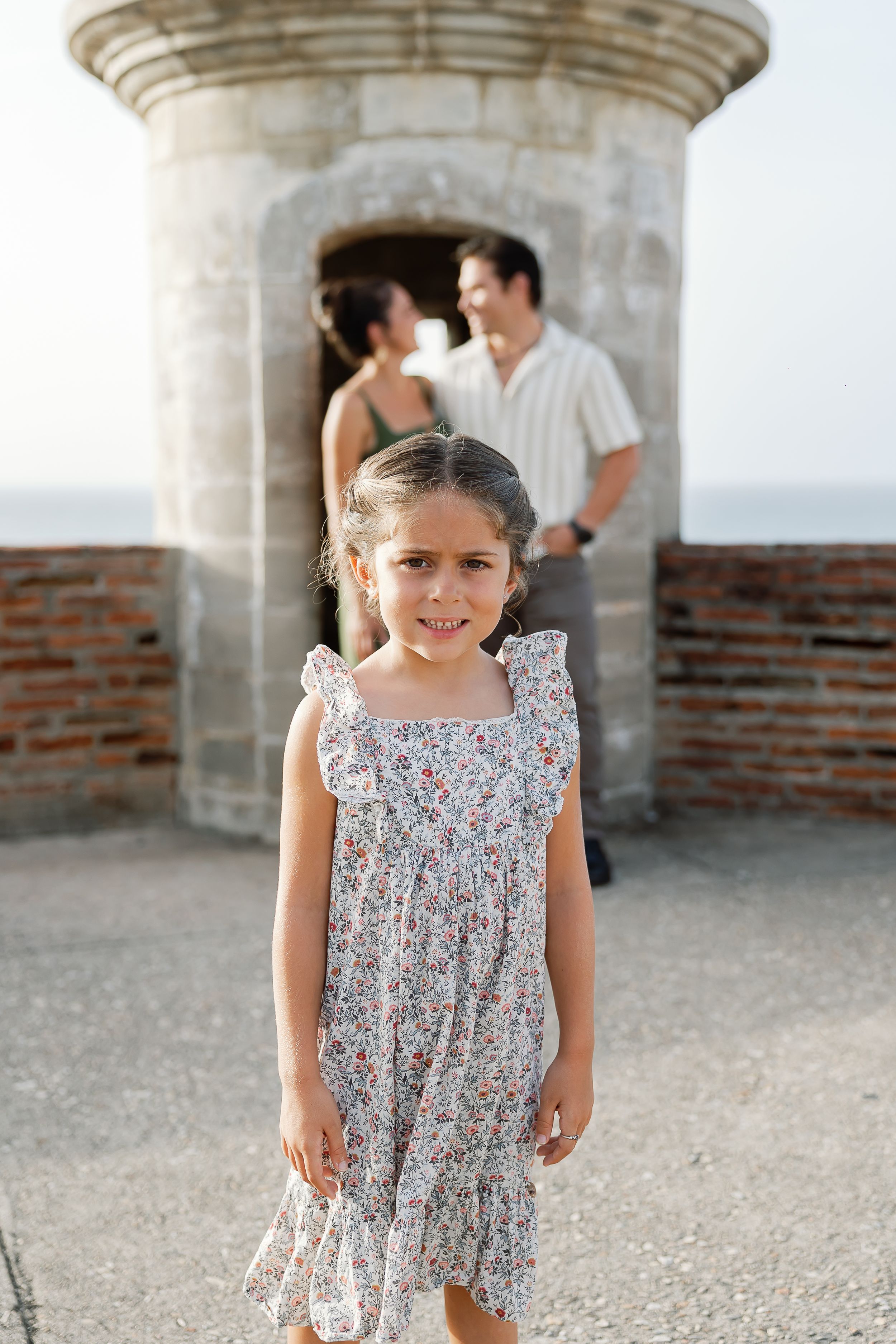 A young girl in a floral dress stands in the foreground, with a couple embracing in the background in front of a historic building.