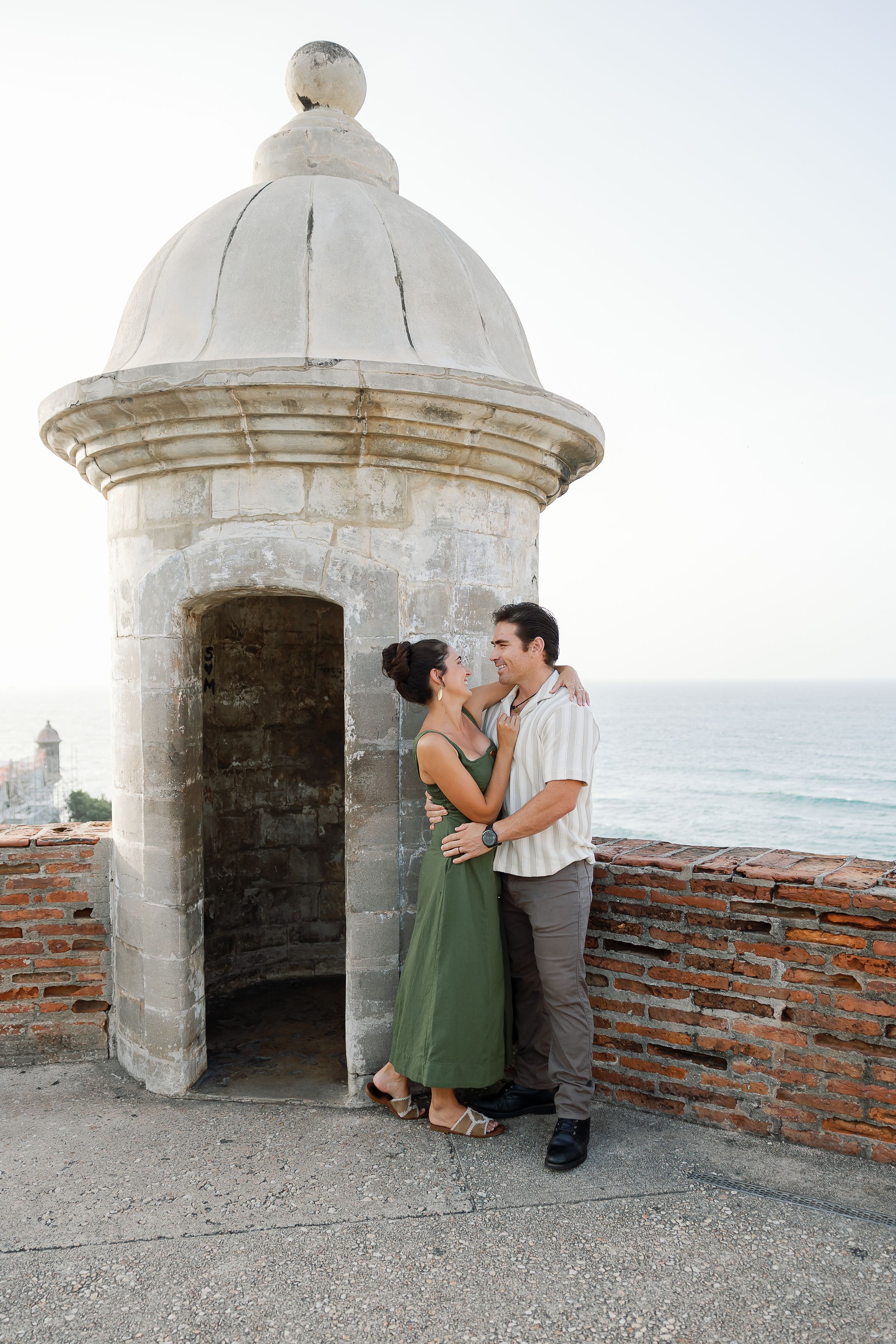 A couple embracing in front of a historic domed building overlooking the ocean.