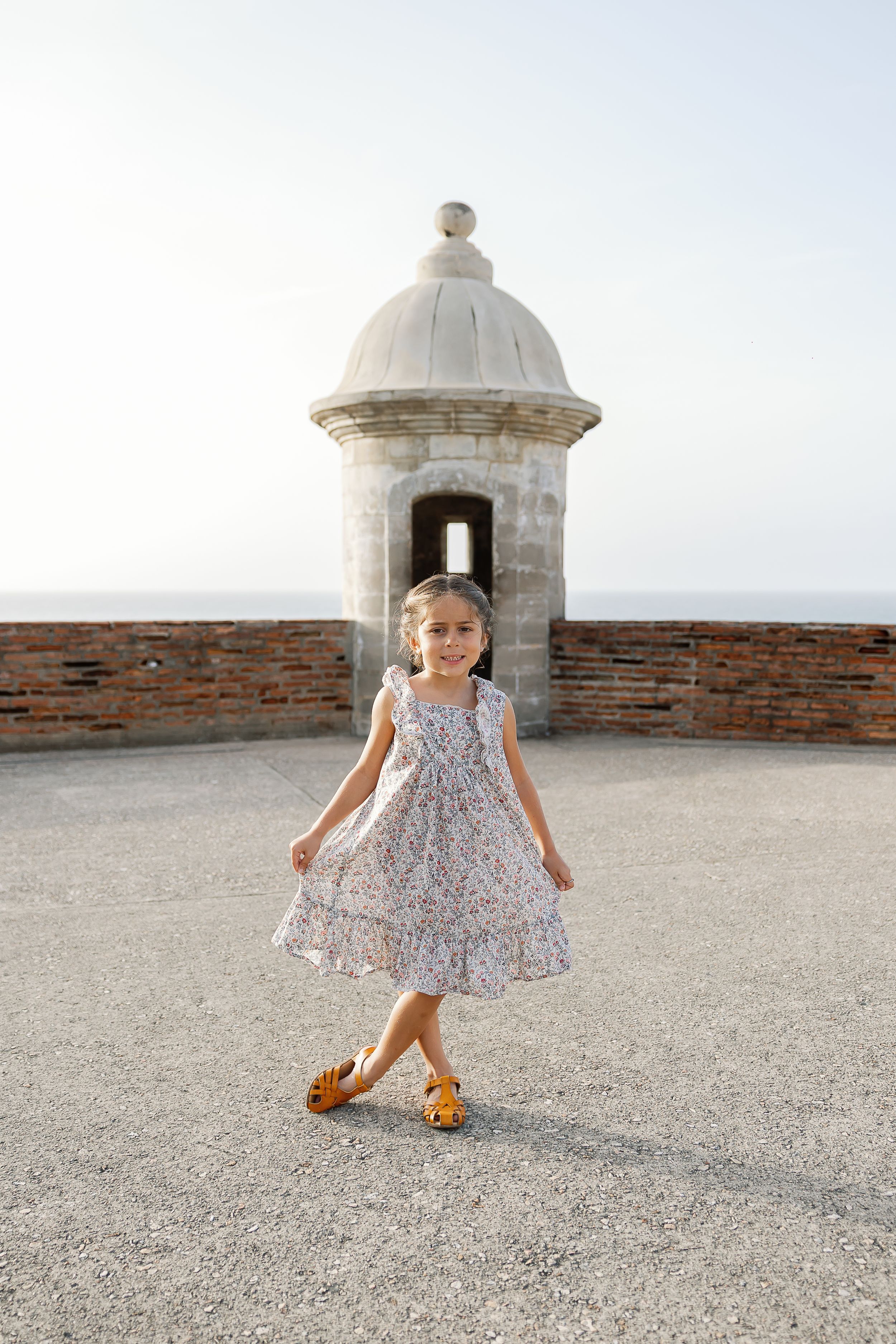 A young girl in a floral dress stands in the foreground in front of a historic domed building overlooking the ocean.