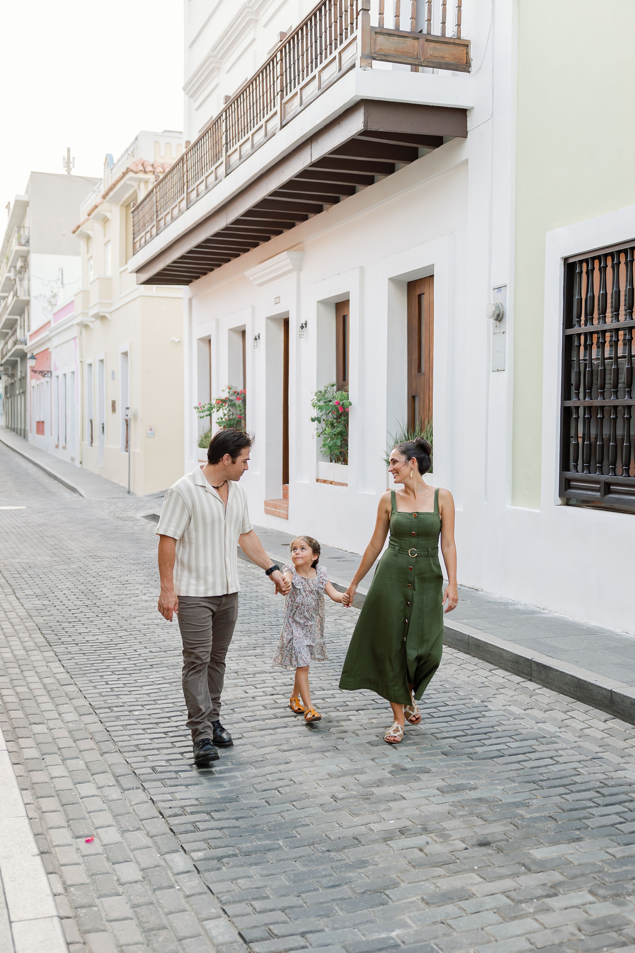 A family of three, a mother, father, and young child, walking together on a cobblestone street in front of historic buildings.