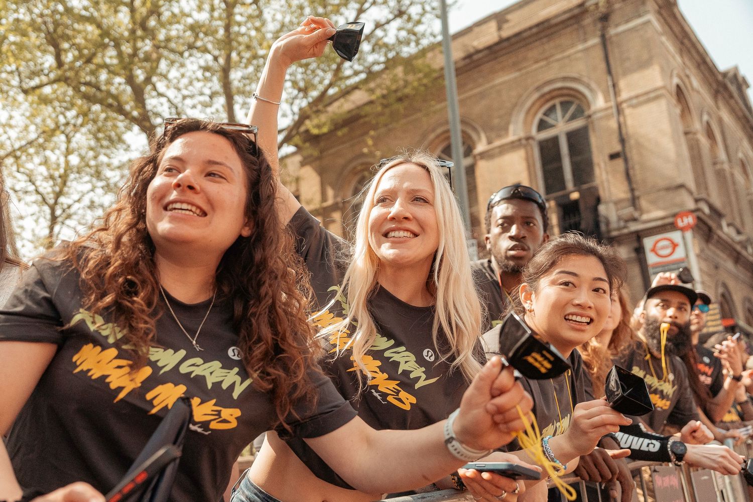 A cheerful crowd wearing matching t-shirts waves and celebrates together outdoors near a historic building.
