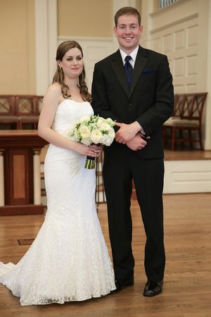 A couple in wedding attire poses in an elegant church interior, with the bride holding a white and green bouquet.