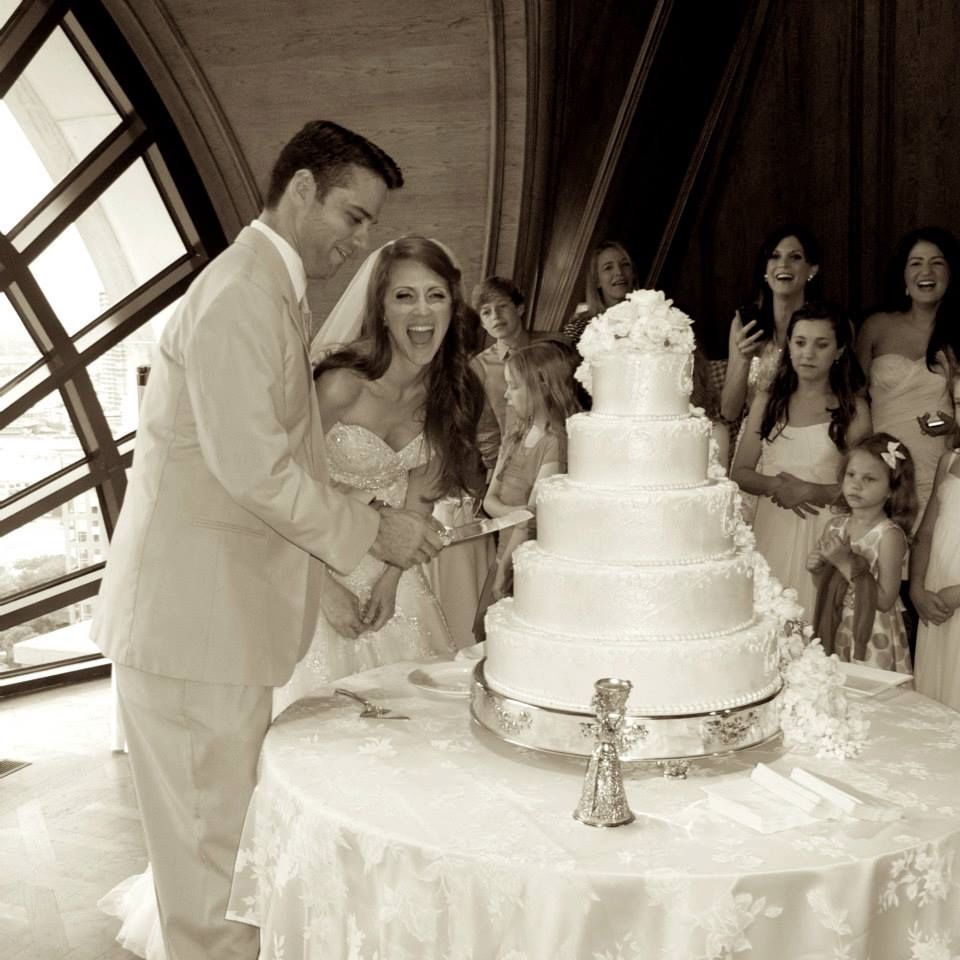 A couple cuts their elegant white tiered wedding cake while guests gather around to watch the traditional moment.