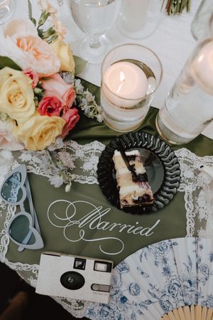 Elegant wedding table with sage green married sign, floral bouquet, candles, disposable camera, and cake slice.