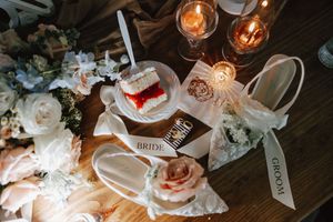 Elegant wedding flat lay with bridal shoes, groom ribbon, ring pillow, roses, and candles on rustic wood table.