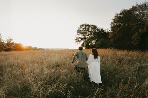 Couple holds hands while walking through golden sunlit field during sunset.