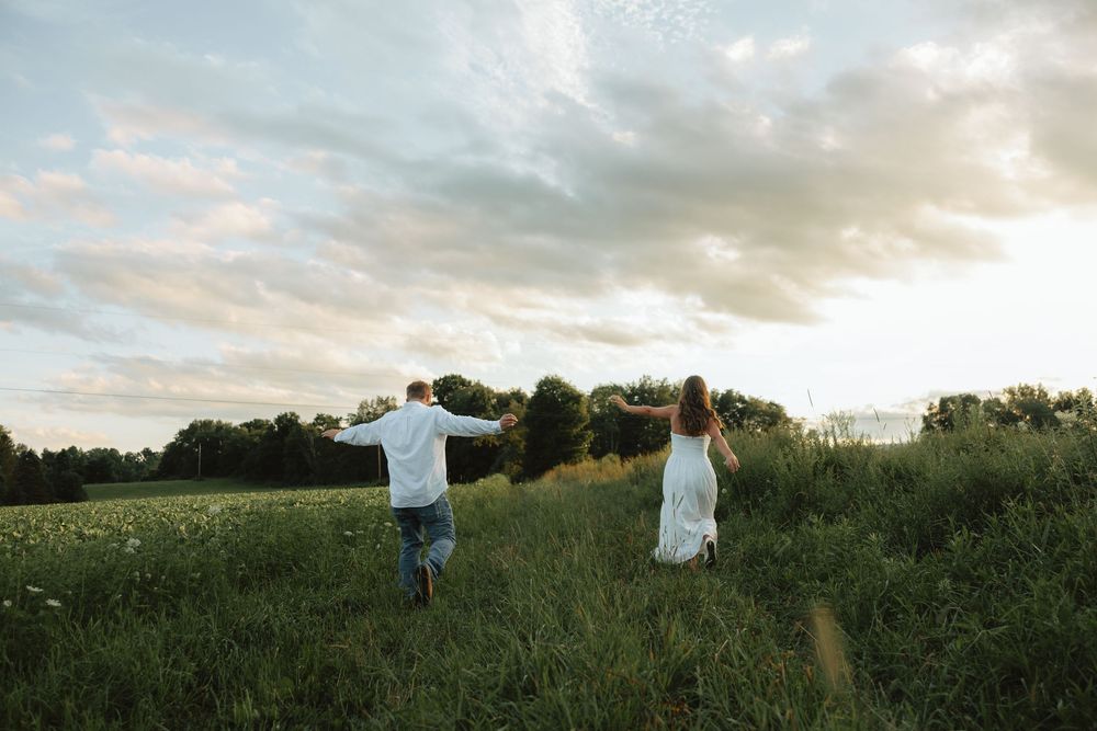 A couple runs hand-in-hand through a lush green meadow at sunset under dramatic cloudy skies.