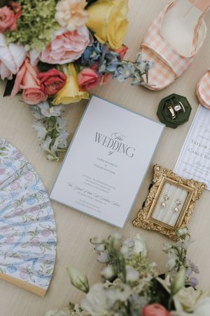 Wedding flat lay with floral bouquet, invitation suite, pearl earrings, gold frame, ring box, and gingham heels.