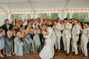 A group of wedding guests dance in celebration under a white tent with string lights during a reception party.