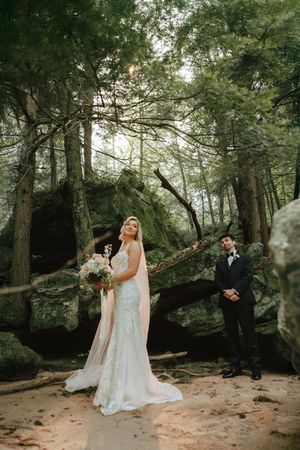 Bride in lace gown with pink veil holds bouquet in forest, groom in tuxedo stands among mossy rocks and tall trees.