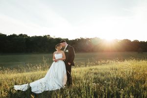 Bride and groom share a romantic kiss in a sunlit meadow with golden hour light and lush green trees in the background.