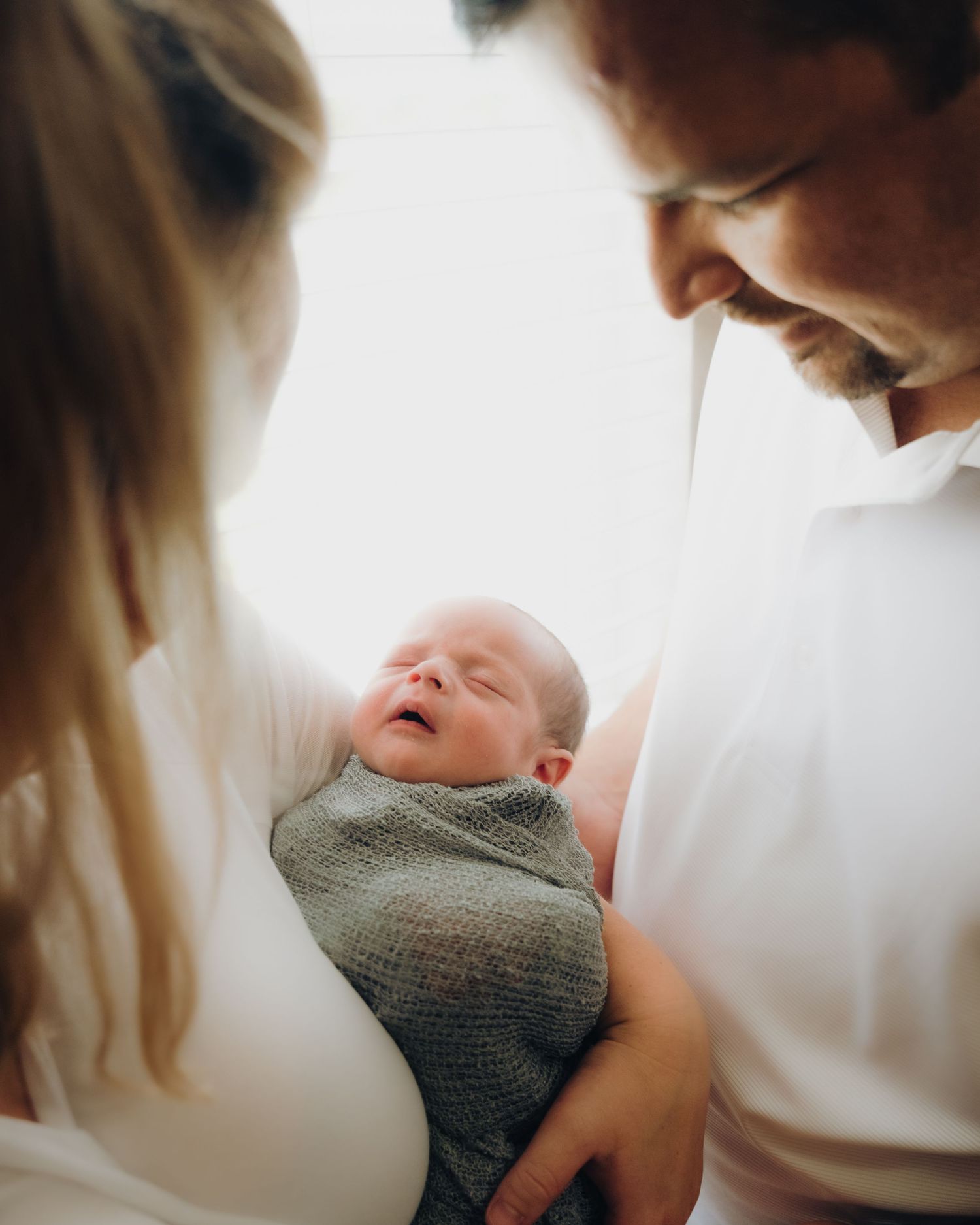 A newborn baby wrapped in gray blanket smiles while being held by their parents.