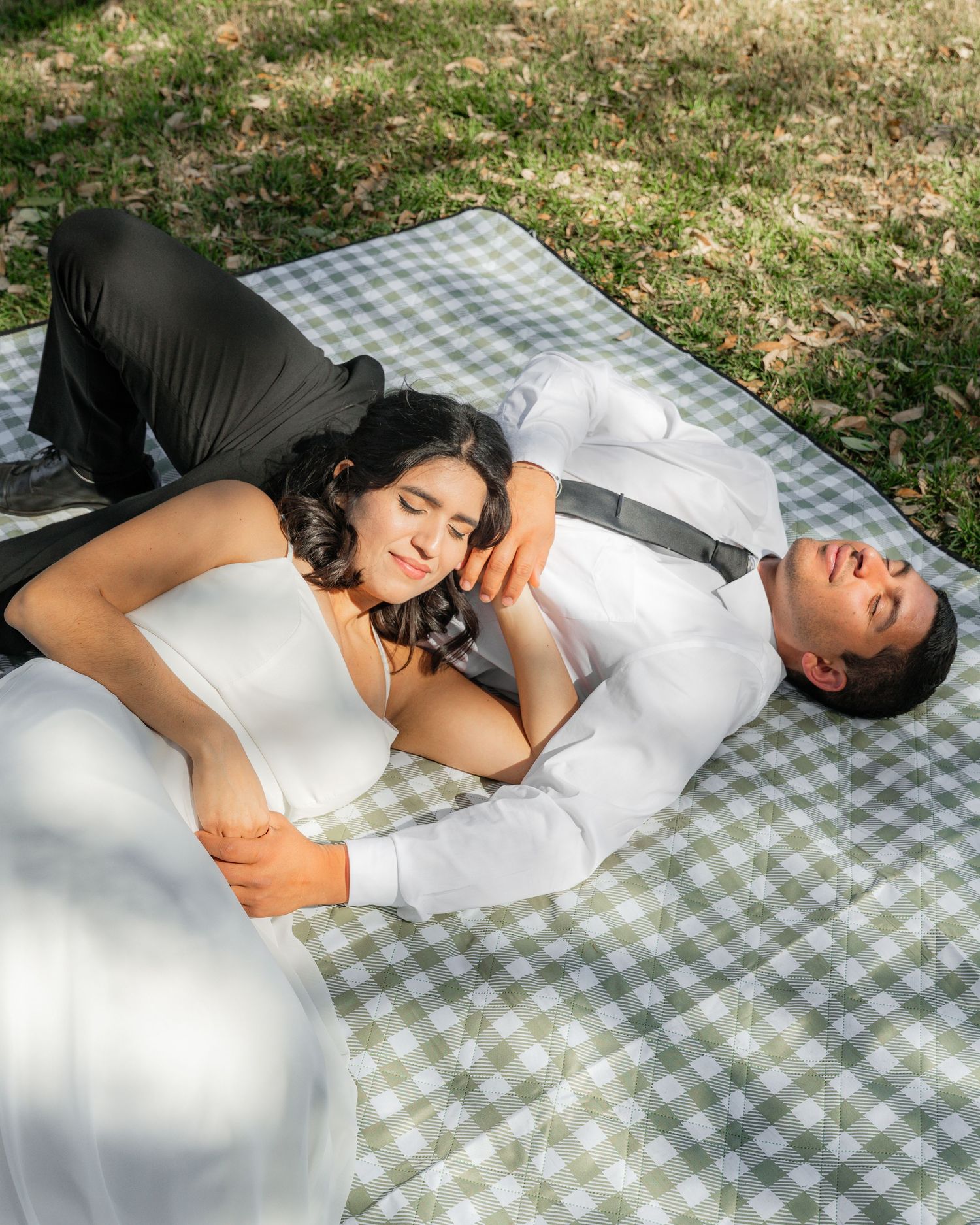 Couple lying on checkered picnic blanket in formal wear during sunny outdoor photoshoot.