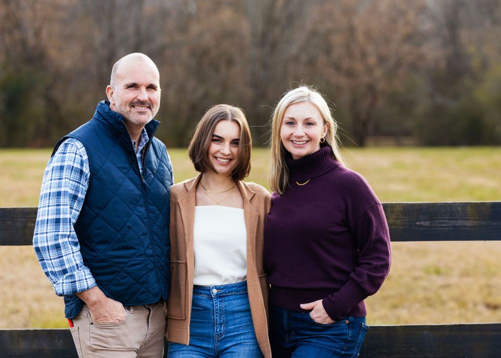 Three people stand together smiling in front of a wooden fence on a fall day wearing casual sweaters and vests.