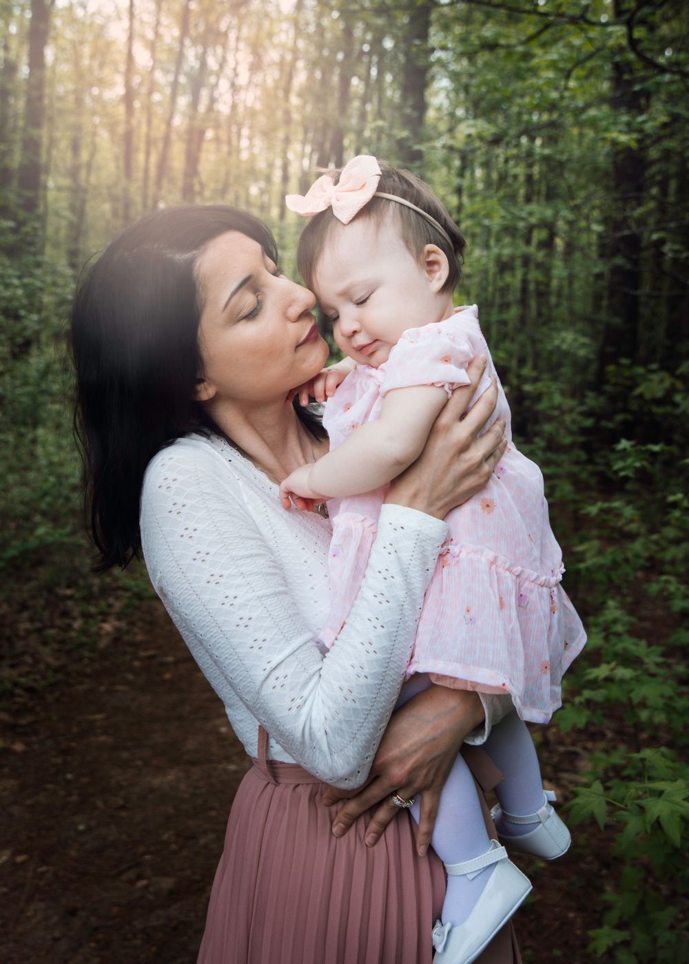 A tender moment shared between mother and baby in a sunlit forest, wearing white and pink outfits.