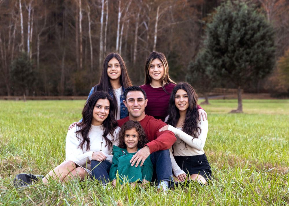 Family portrait of six people sitting together on grass with trees in the background.