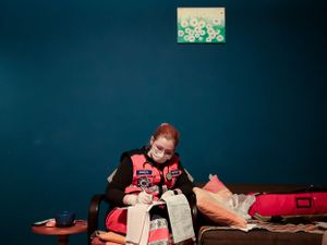 Person sitting on floor sorting through clothing and items against dark teal wall with small floral artwork.