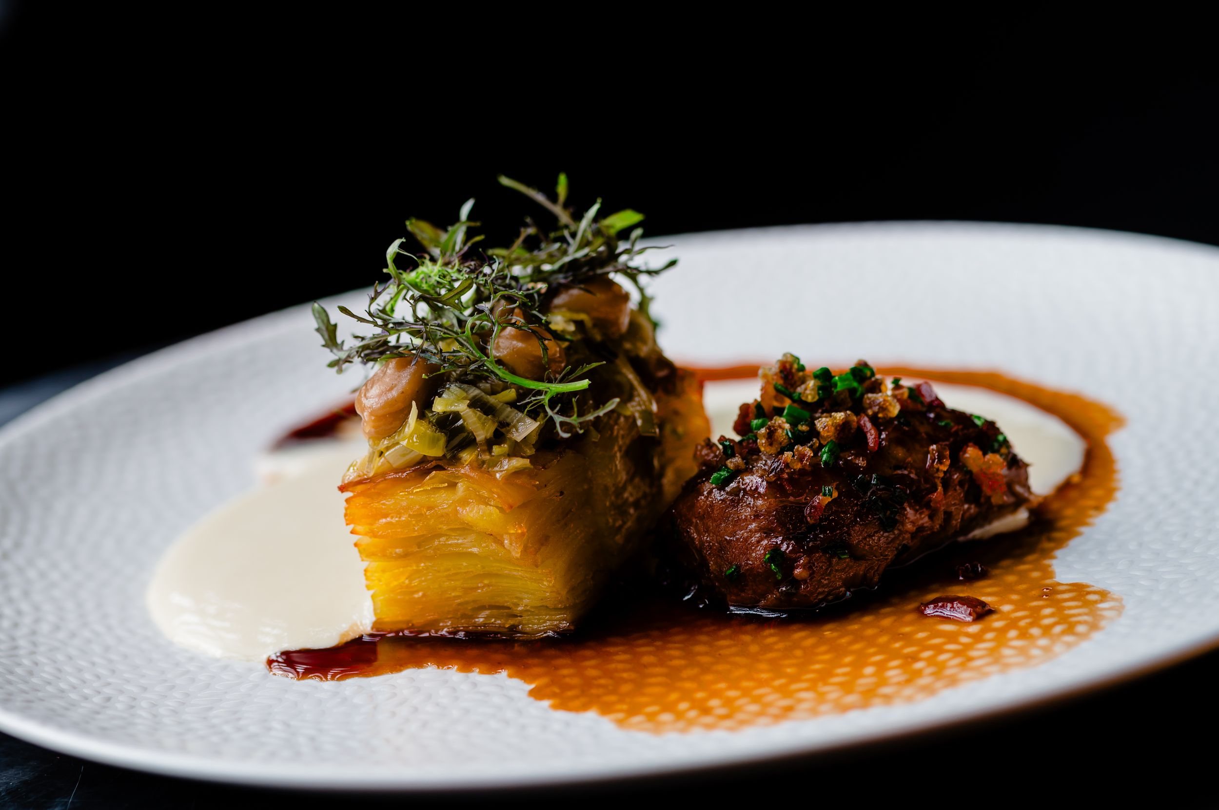 Gourmet plated dish featuring braised meat and roasted potatoes with microgreens garnish in a rich sauce.