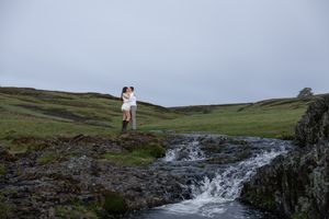 Couple kissing on rocky outcrop beside waterfall in lush green landscape.
