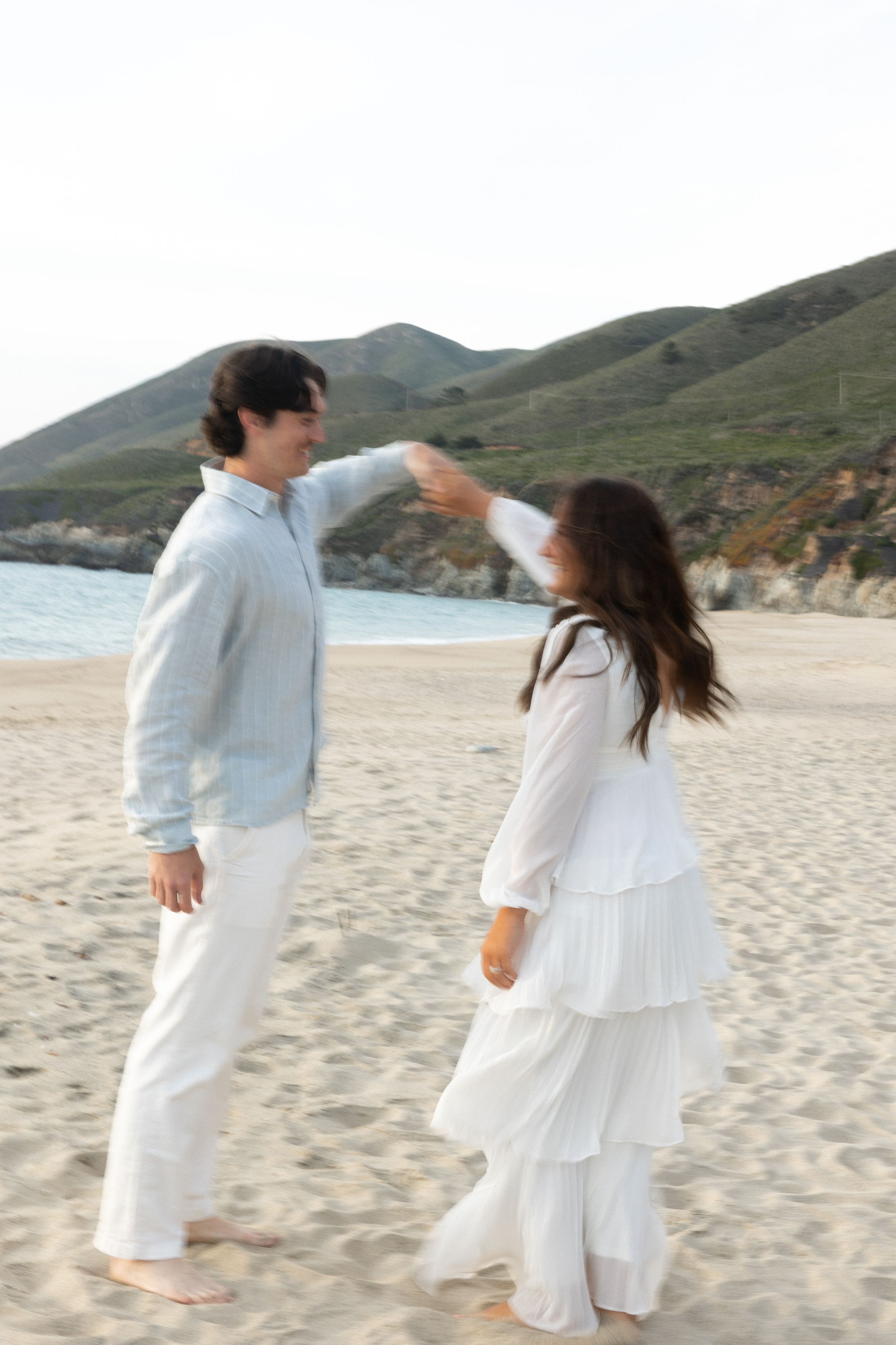 Couple in white outfits dancing on a sandy beach with green hills and ocean in the background.