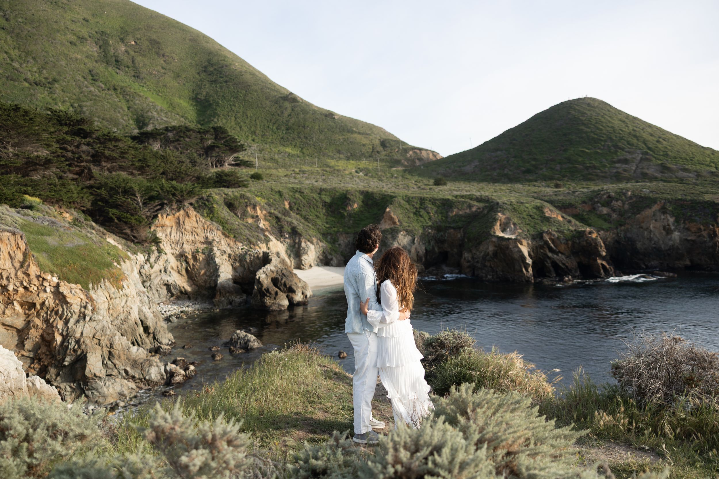 Couple in white attire standing on coastal cliffs overlooking a scenic cove with green hills in Big Sur, California.