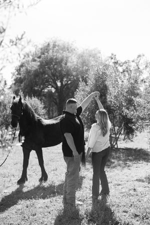 Black and white photo of a couple dancing near a horse in a sunlit outdoor setting with trees in the background.