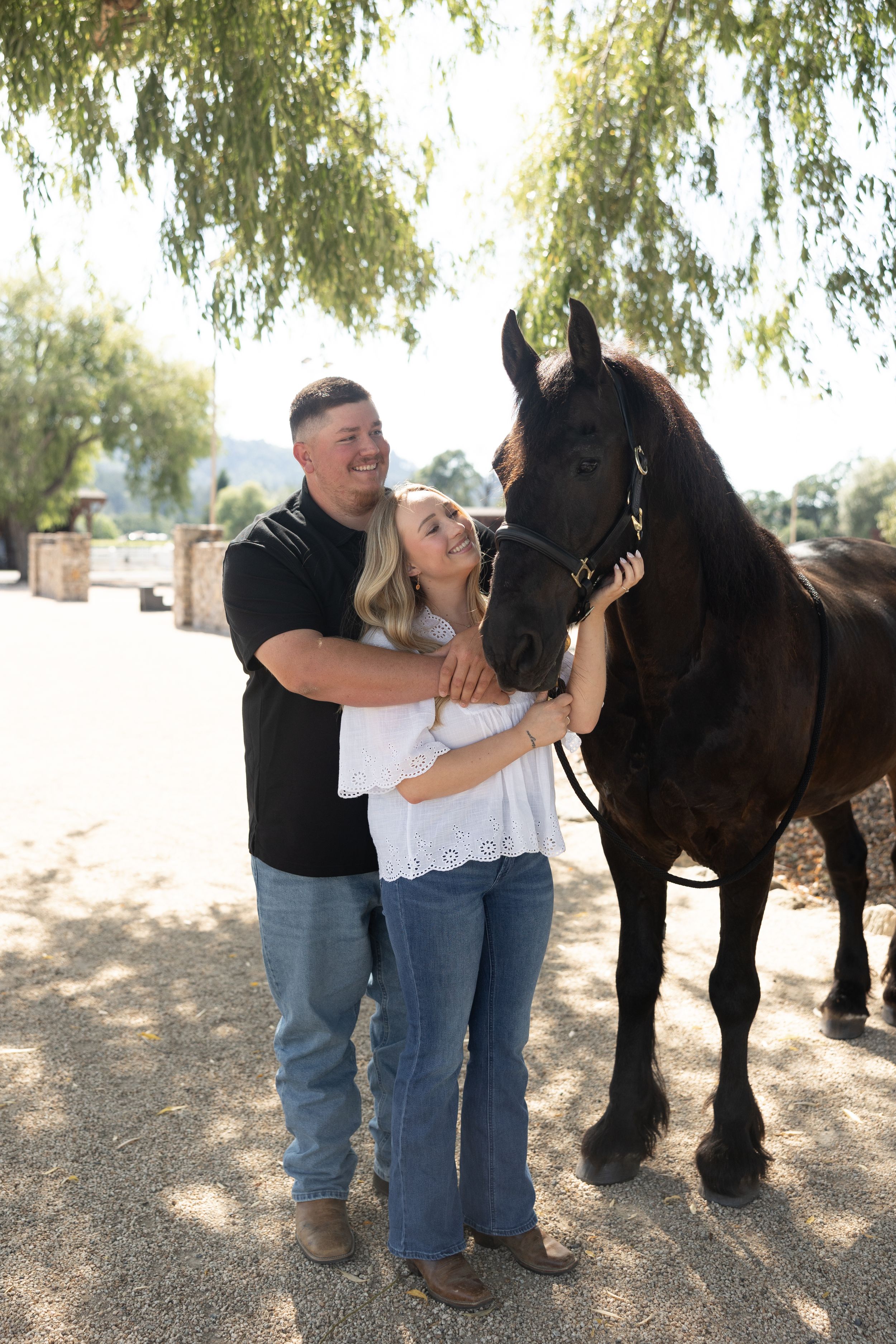 Couple smiling and embracing beside a dark brown horse outdoors under a willow tree on a sunny day.