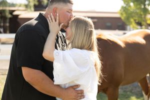 Couple sharing a romantic kiss outdoors near a brown horse on a sunny farm day.
