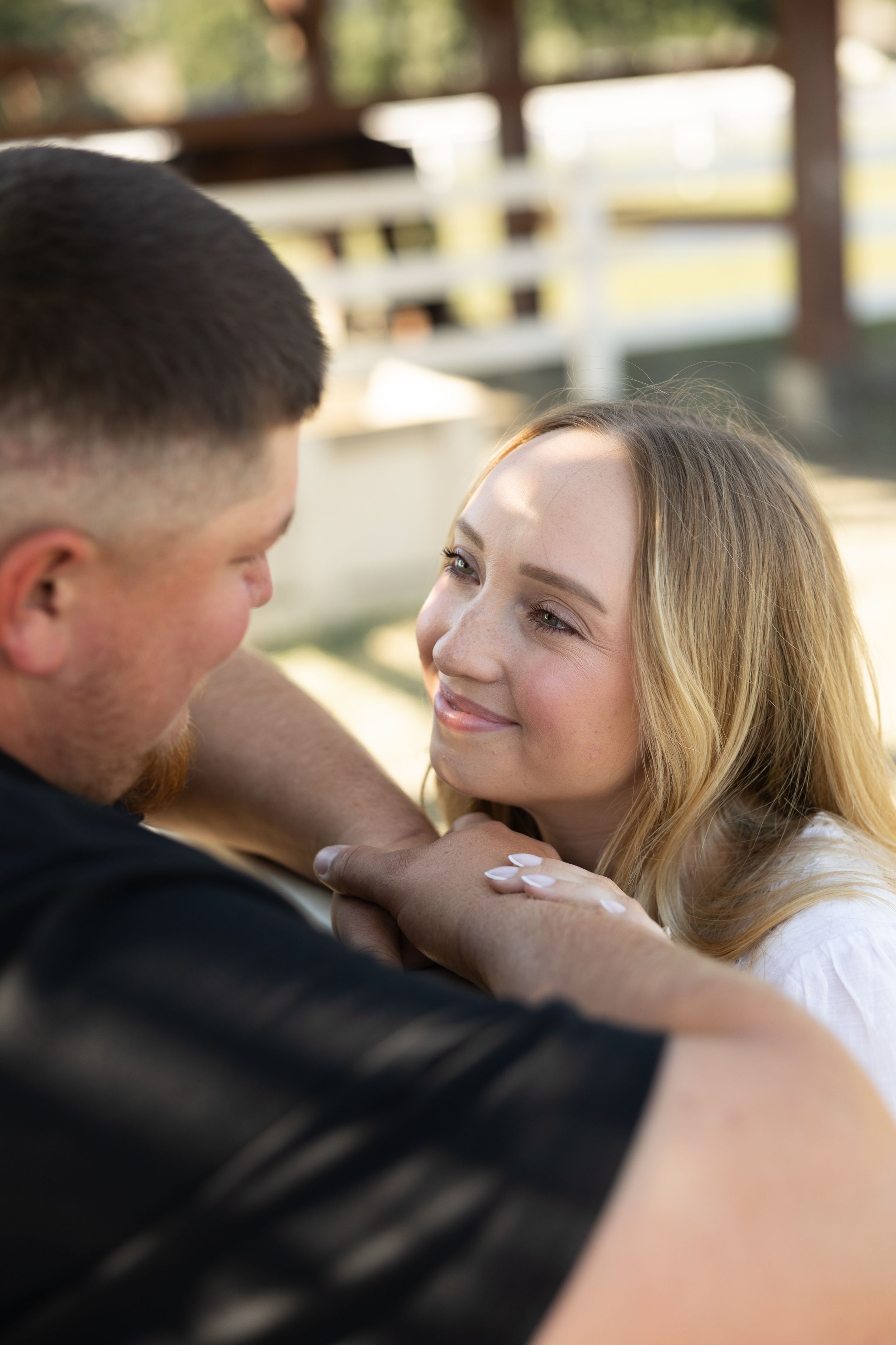 Young woman smiling lovingly at a man while resting her hands on his chest outdoors near a white fence.