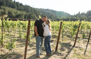 Couple sharing a romantic moment kissing in a lush vineyard with green grapevines and forested hills in background.