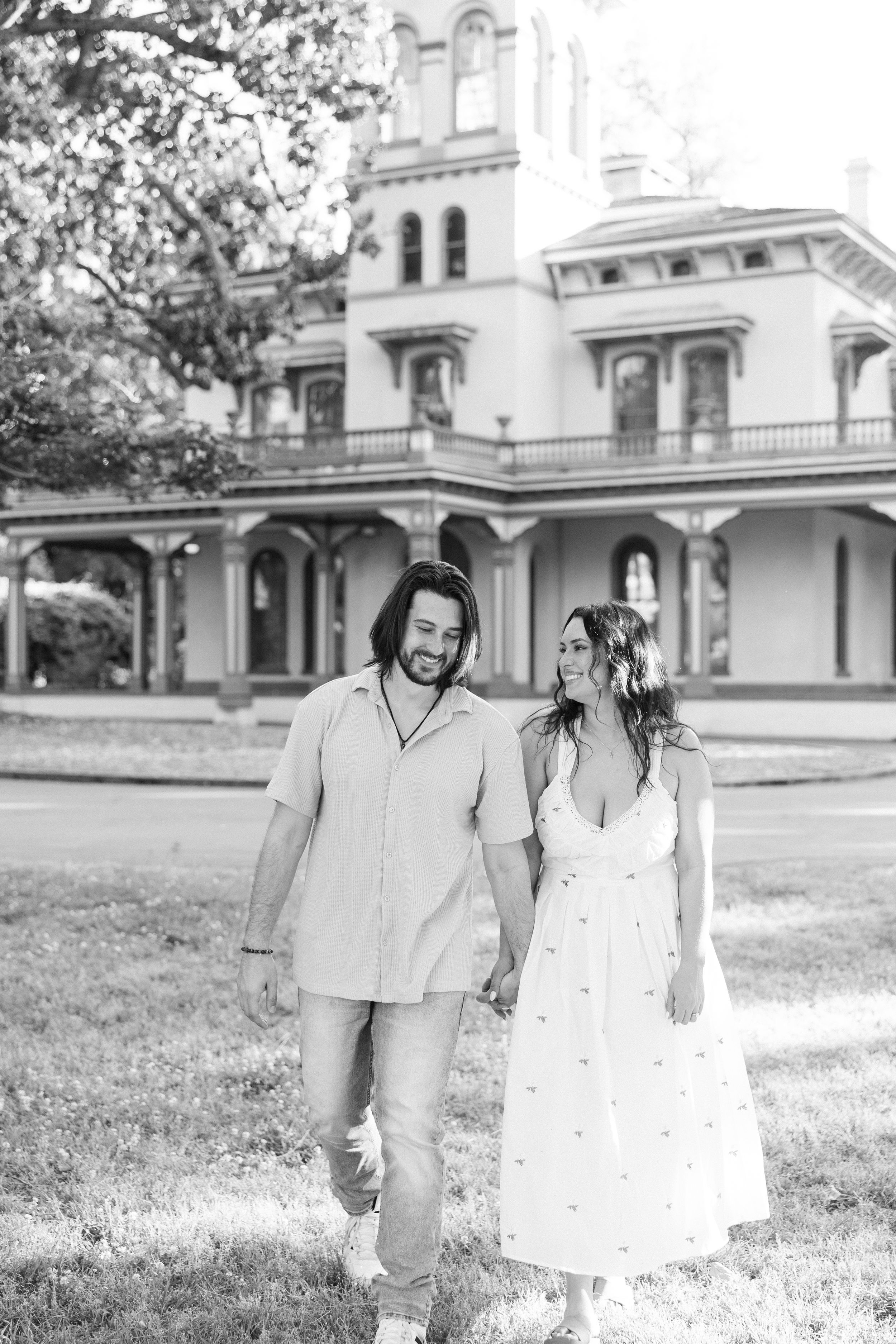 Smiling couple in black and white photo walking hand in hand by historic building.