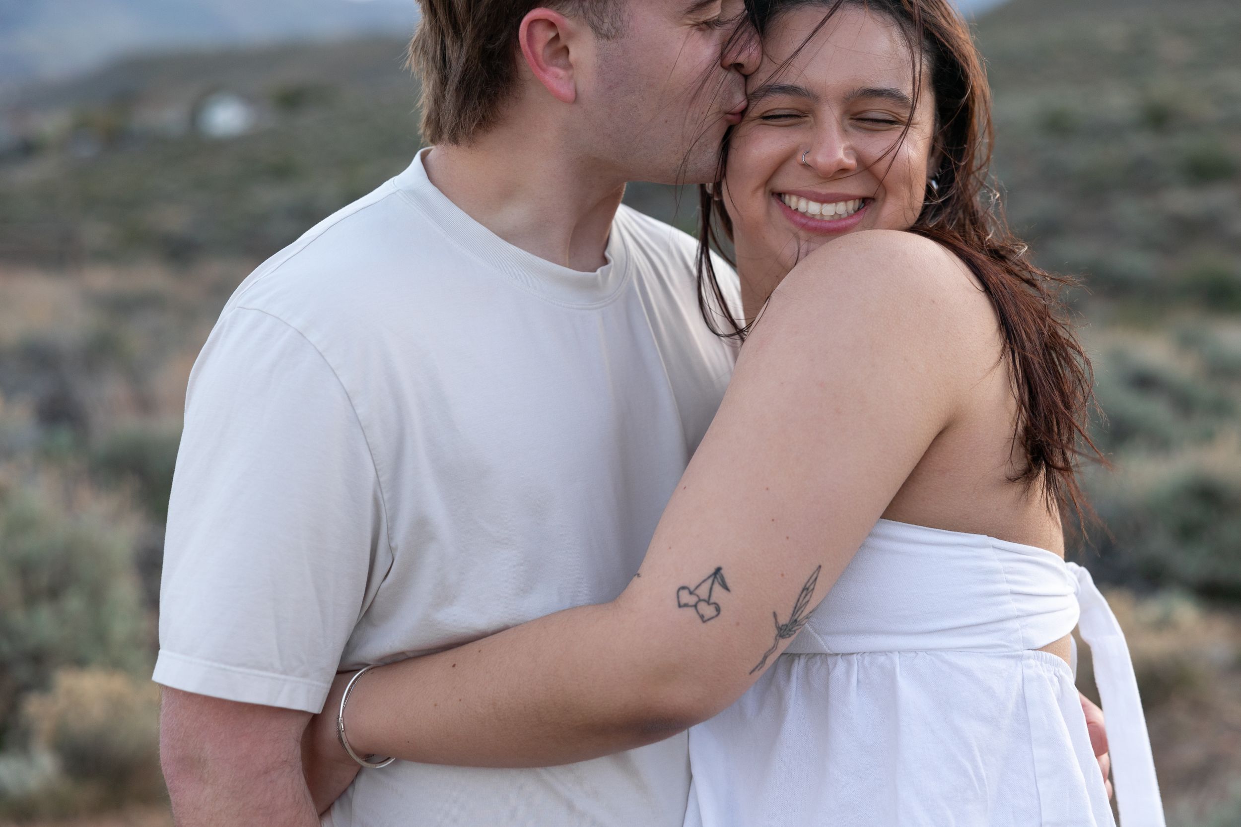 Man kissing smiling woman with floral tattoo outdoors in desert setting.