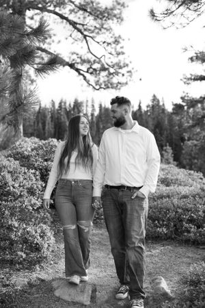 Black and white photo of smiling couple walking hand in hand along a pine tree lined mountain path.