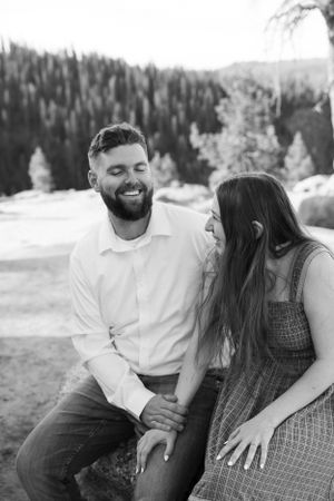 Black and white photo of laughing couple sitting together outdoors with snowy pine forest in the background.