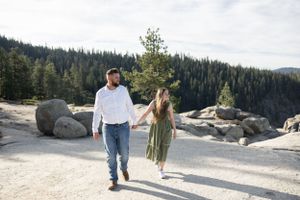 Couple walking hand in hand on a sunny granite overlook with pine forest backdrop.