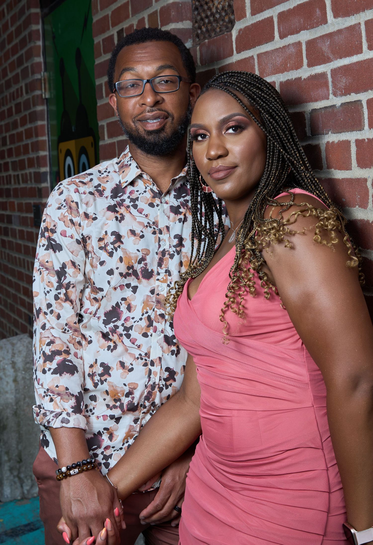 Couple poses together against brick wall backdrop in coordinating pink and floral outfits for engagement photos.