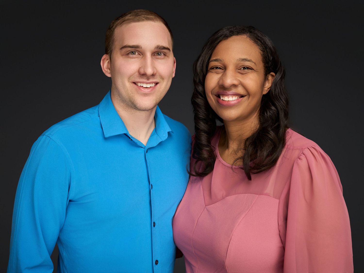 Professional studio portrait of couple in blue shirt and mauve top against black background.