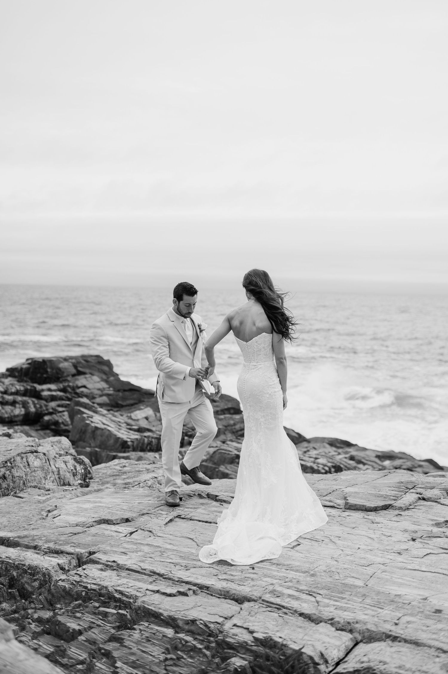 A couple stands on rocky coastal cliffs overlooking the ocean in black and white.