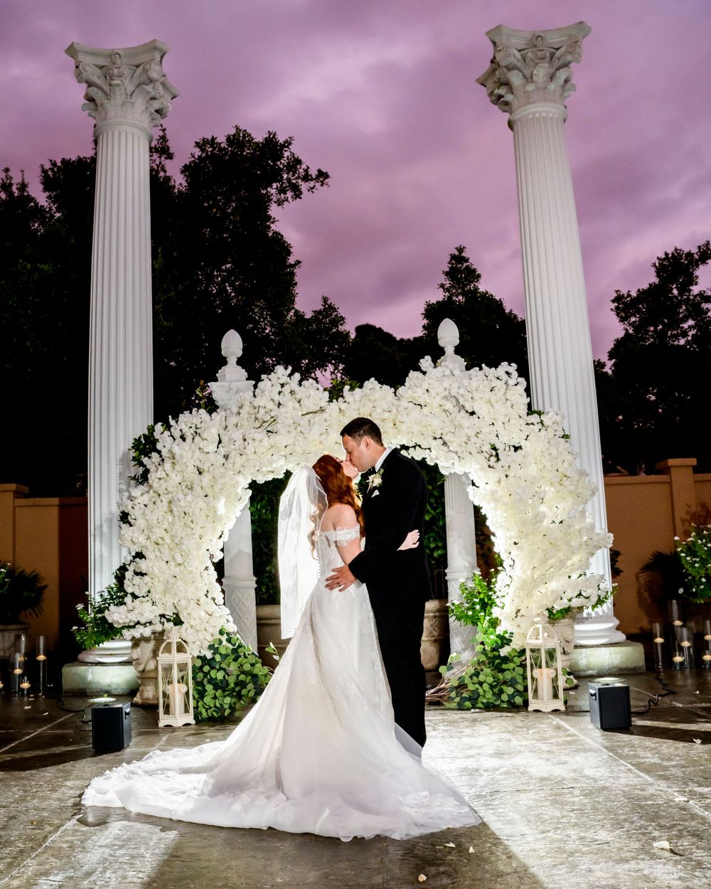 Wedding couple poses under illuminated white columns and floral arches during twilight reception.