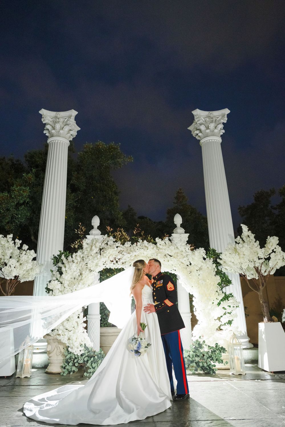 Bride and groom share a romantic moment at night surrounded by elegant white Greek columns and floral decorations.